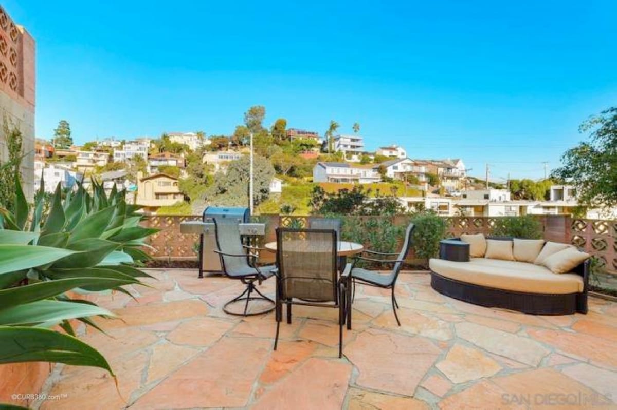 An enclosed terrace features natural stone flooring and a seating area with a round table and four chairs. A circular lounge chair offers a cozy nook, with verdant plants nearby. The backdrop showcases a hillside neighborhood with residential homes and greenery under a clear blue sky.
