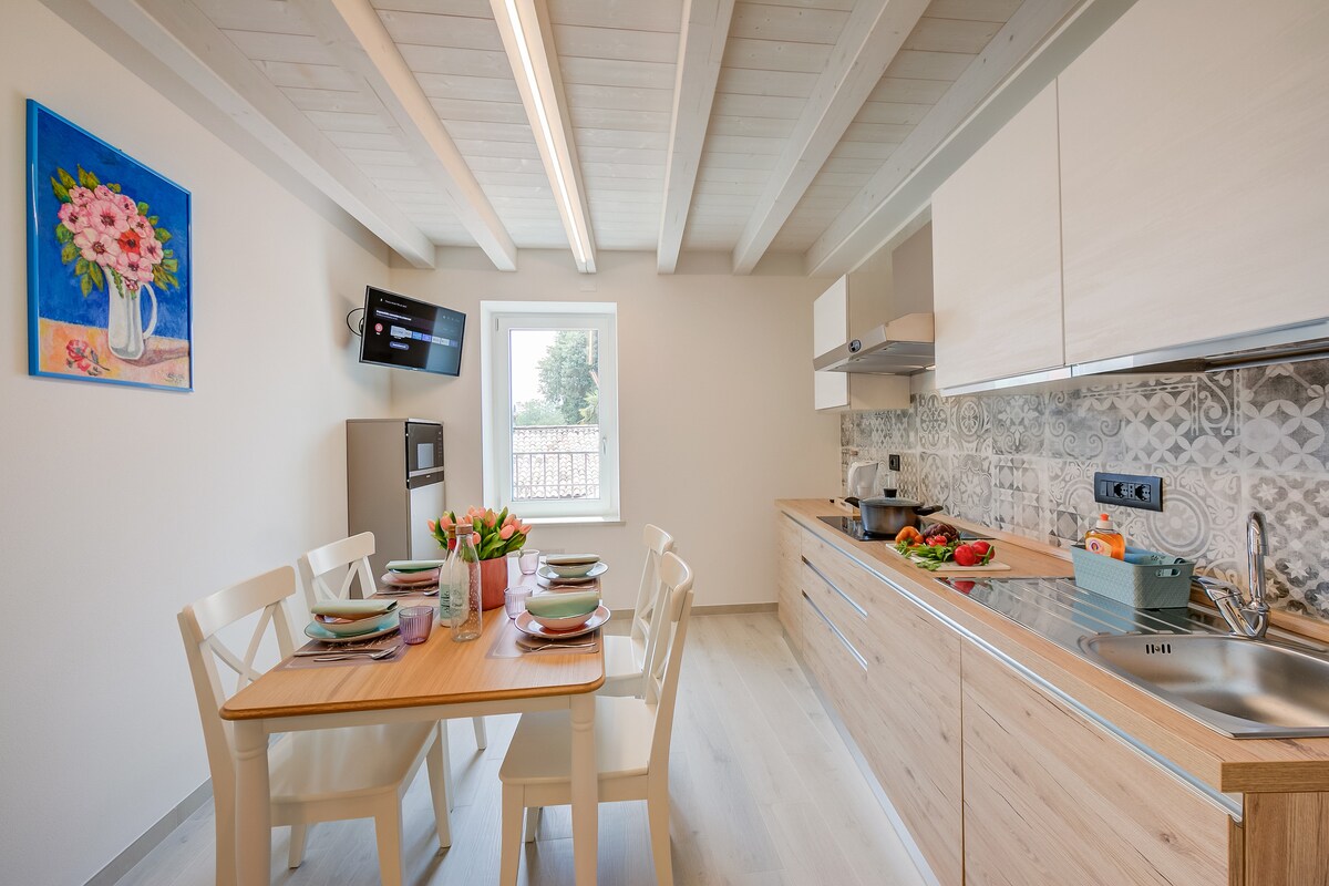 A modern kitchen is equipped with light wood cabinetry and a stainless steel countertop. A dining table is set for four, adorned with vibrant plates and fresh flowers. A flat-screen TV is mounted on the wall, complemented by a window that allows natural light to enter.