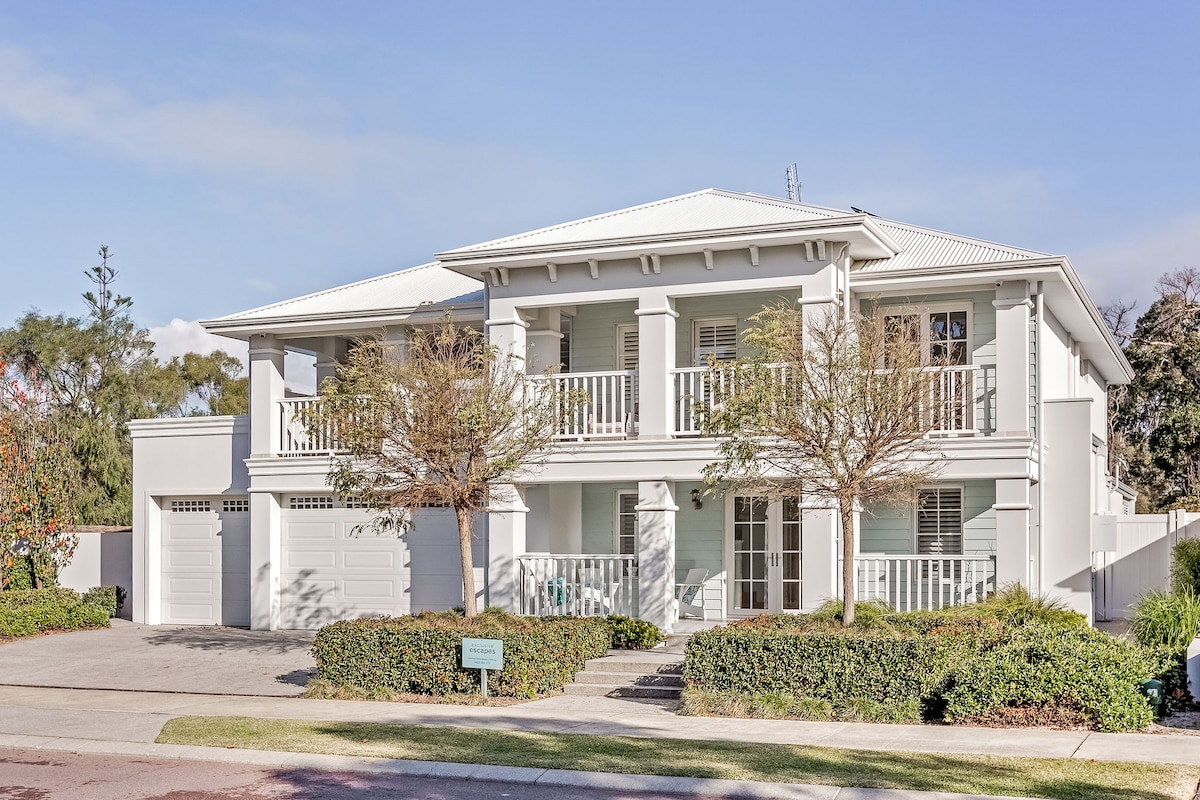 A two-storey home showcases a clean, white façade adorned with balconies on the upper level. Lush greenery and well-maintained landscaping frame the front yard, leading to a neat driveway and garage. Sunlight illuminates the exterior, creating a serene and inviting appearance.