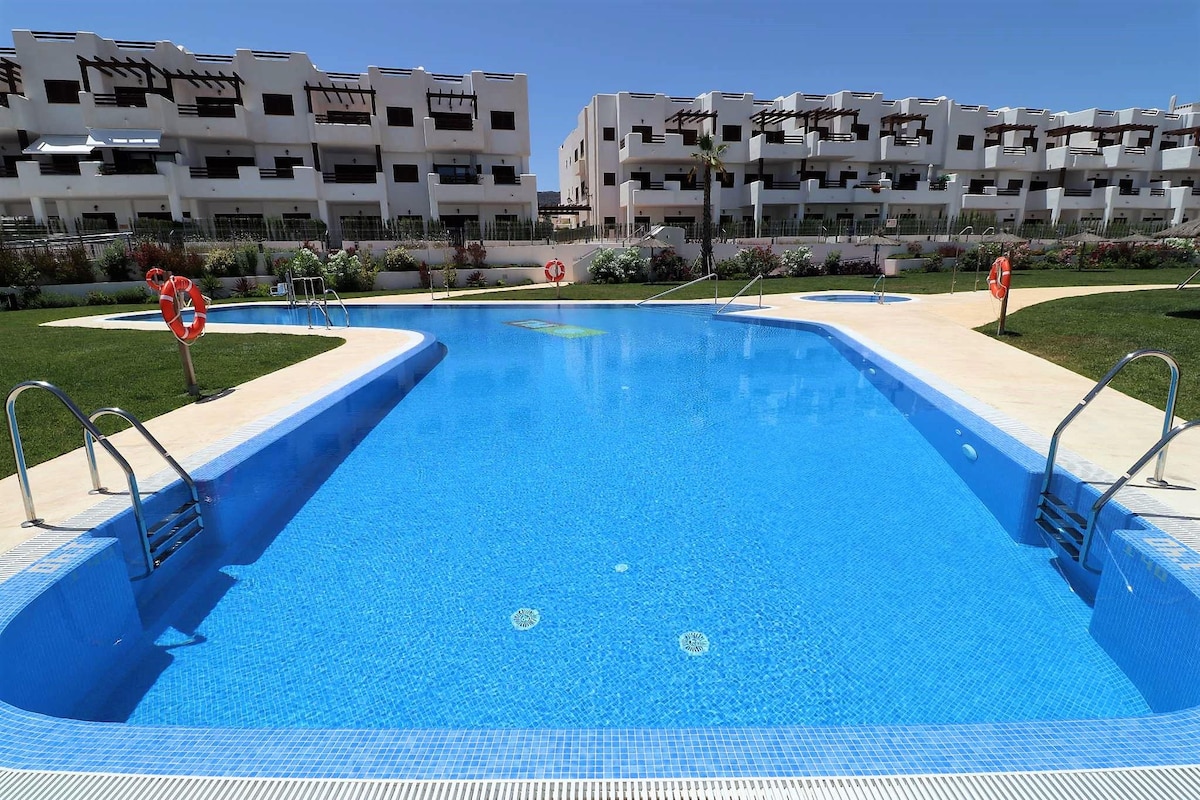 A communal swimming pool is surrounded by well-maintained gardens and modern apartment buildings. The pool features bright blue water and steps for easy access. Lifebuoys are positioned along the edges for safety, enhancing the tranquil atmosphere of the outdoor space.