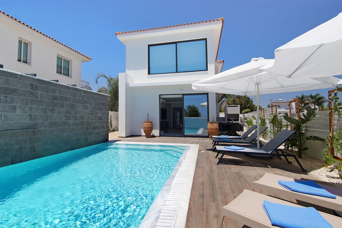 A private swimming pool with clear blue water is surrounded by a wooden deck and comfortable lounge chairs. An umbrella provides shade, while adjacent, a glass door leads to an indoor living space, enhancing the seamless transition between indoor and outdoor areas.