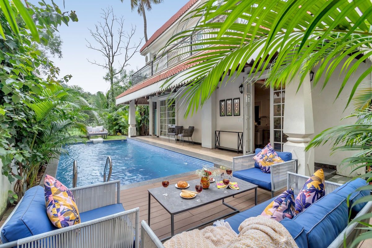 An outdoor area features a private pool surrounded by lush greenery. Comfortable blue seating arrangements with colorful pillows are set around a table adorned with snacks and beverages. The villa's exterior is visible in the background, complemented by tropical plants enhancing the serene environment.