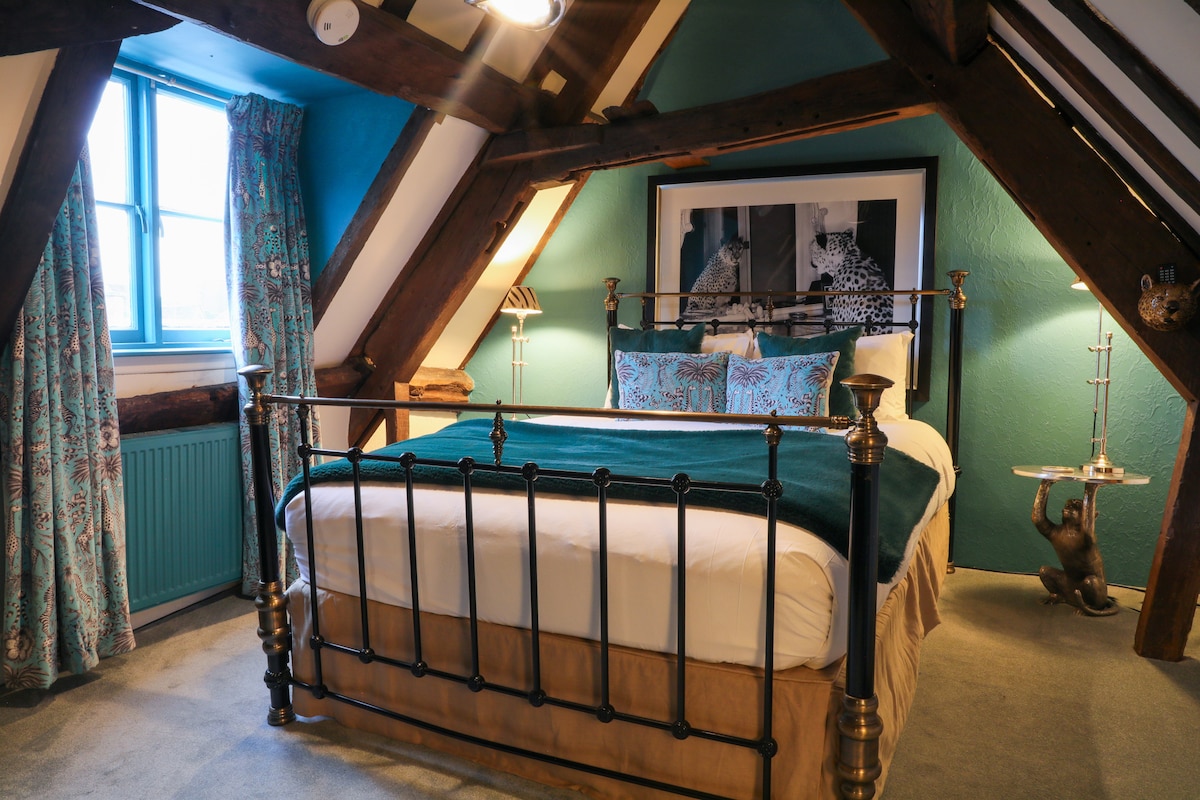 A spacious bedroom is framed by wooden beams and features a brass bed with an elegant headboard. Colorful cushions complement the bedding. A large window offers natural light, draped with patterned curtains. Stylish bedside lamps and framed artwork add to the room's charm.