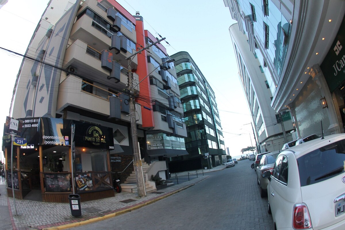The image depicts a street view featuring modern buildings flanking either side. A commercial space with outdoor seating is visible on the left, while a tall glass structure is on the right. The street is lined with parked vehicles under a clear sky.