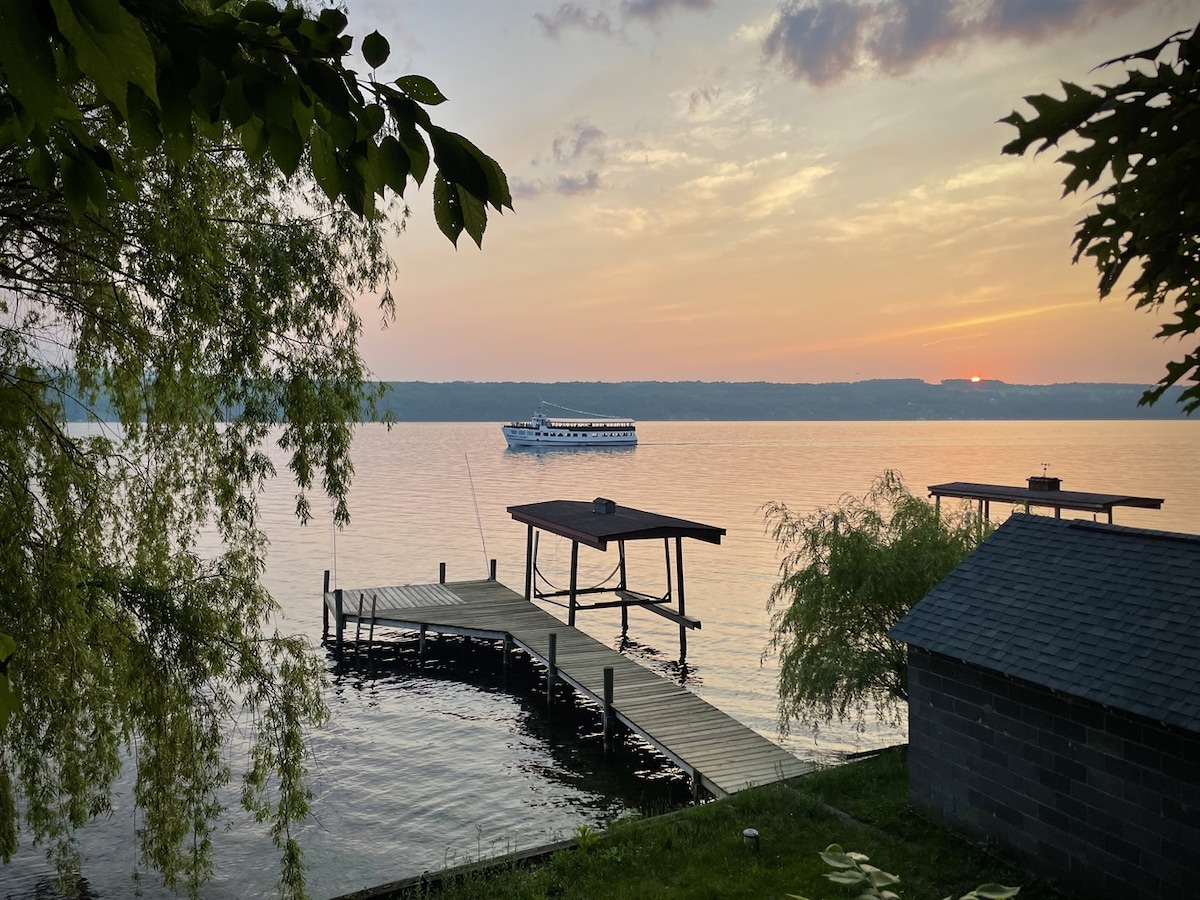 The serene waterfront scene captures a boat gently gliding across Seneca Lake at sunset, with the sky displaying soft hues of orange and purple. A dock and boathouse are visible in the foreground, surrounded by lush greenery and the calm water.