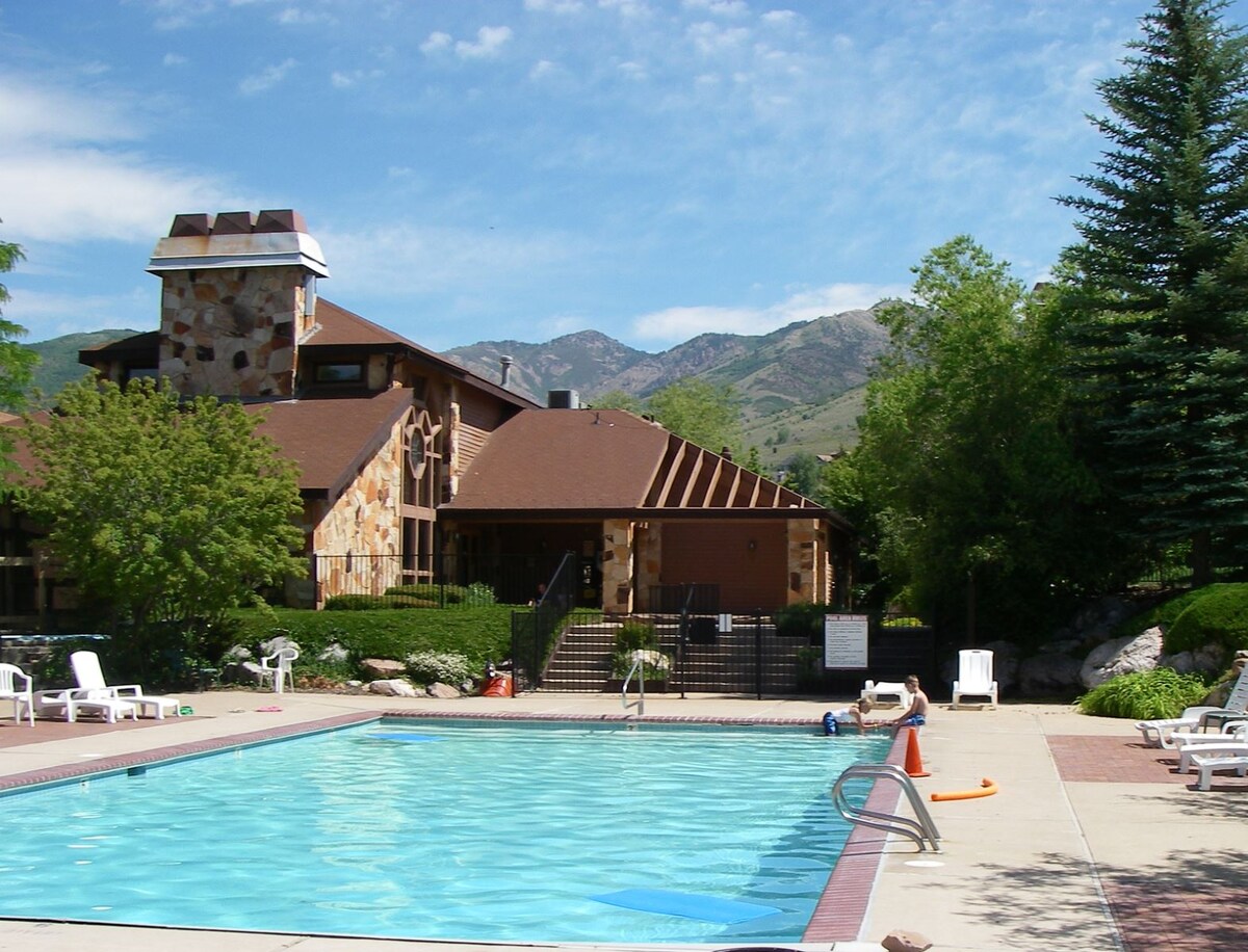 The outdoor pool is surrounded by a spacious deck, featuring several lounge chairs for relaxation. A multi-story stone building with a wooden roof structure is visible in the background, framed by greenery and mountains, enhancing the serene environment.