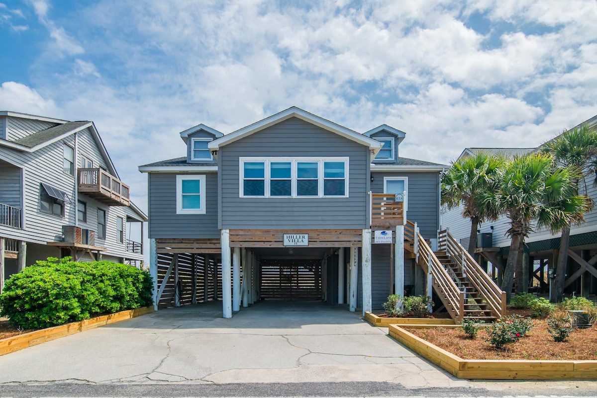 The exterior of the oceanfront home showcases a grey facade with large windows, allowing for ample natural light. Palm trees and landscaped areas provide greenery at the front. Stairs lead to the main entrance, and covered parking is available underneath the house.