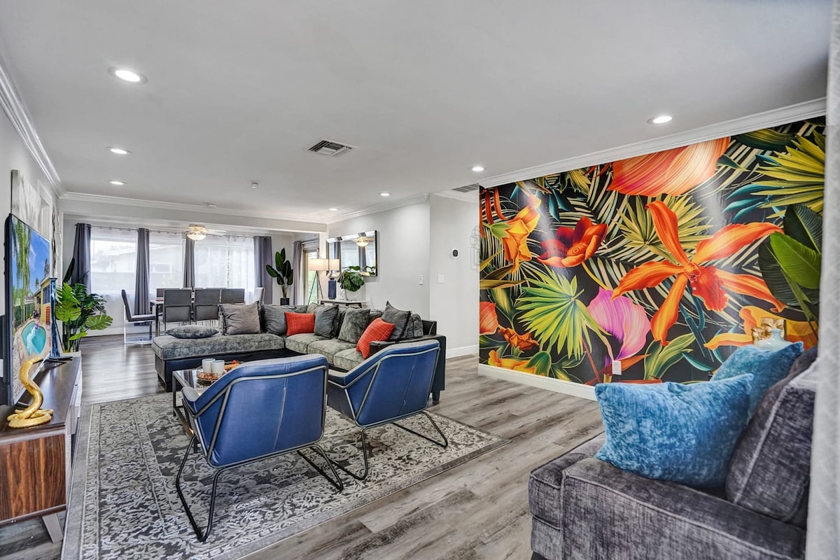 An open living area features comfortable sectional sofas and armchairs arranged around a coffee table. A vibrant floral mural adds a pop of color to the wall, while natural light brightens the space through large windows. The area is designed for relaxation and socializing.