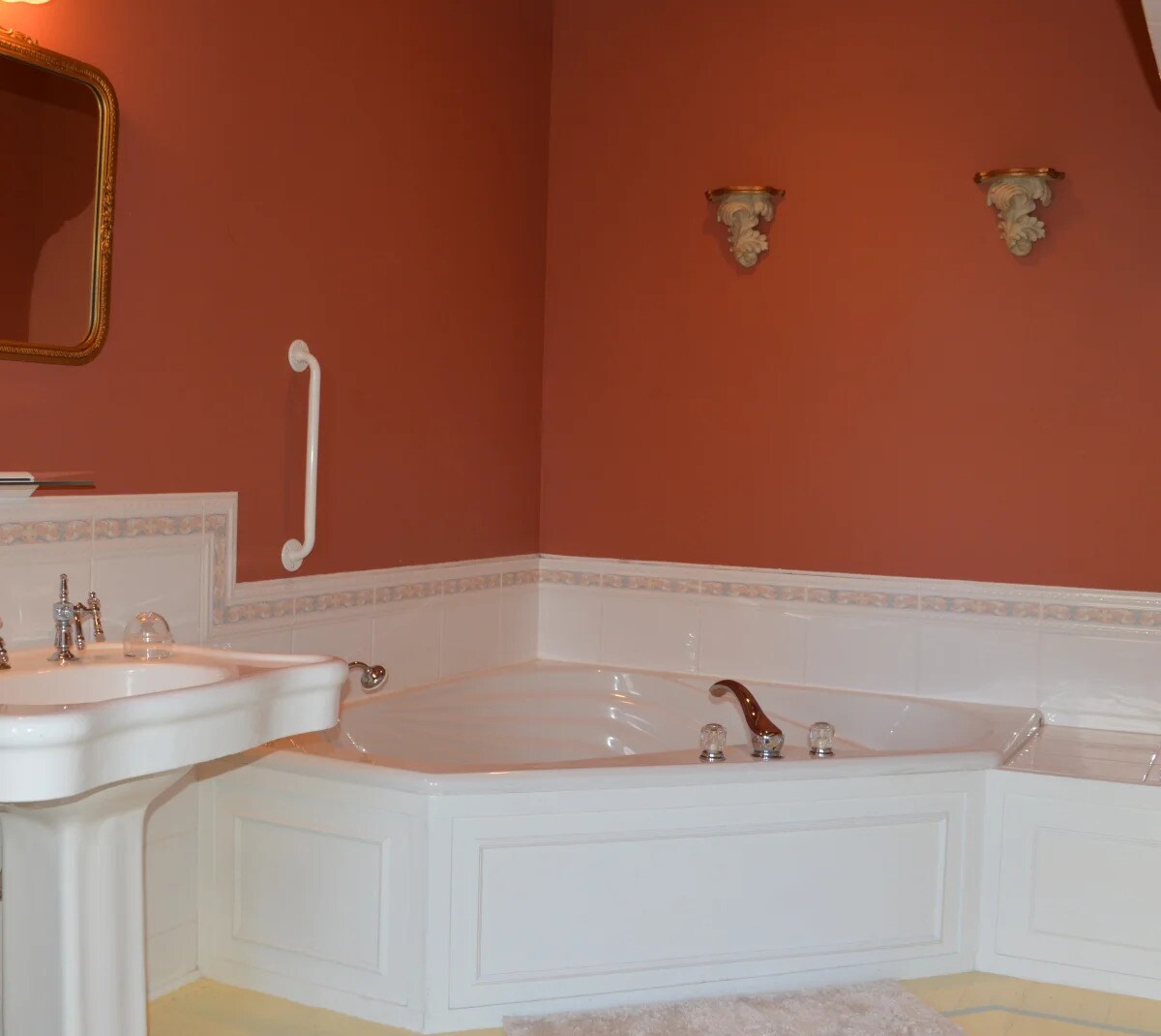 A whirlpool tub is positioned in the corner of a warm-toned bathroom, with a large mirror above a vanity sink. Accents of elegant wall sconces complement the soothing atmosphere, while a plush rug is placed on the floor nearby.