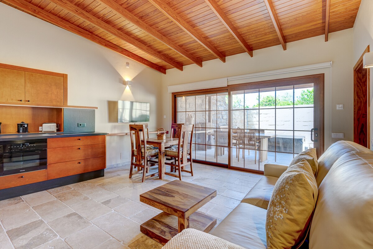 The spacious kitchen-living room features warm wooden ceilings and tasteful cabinetry. A cozy leather sofa sits next to a wooden coffee table. A dining area with a table and four chairs is positioned near large sliding doors, leading to an outdoor terrace.