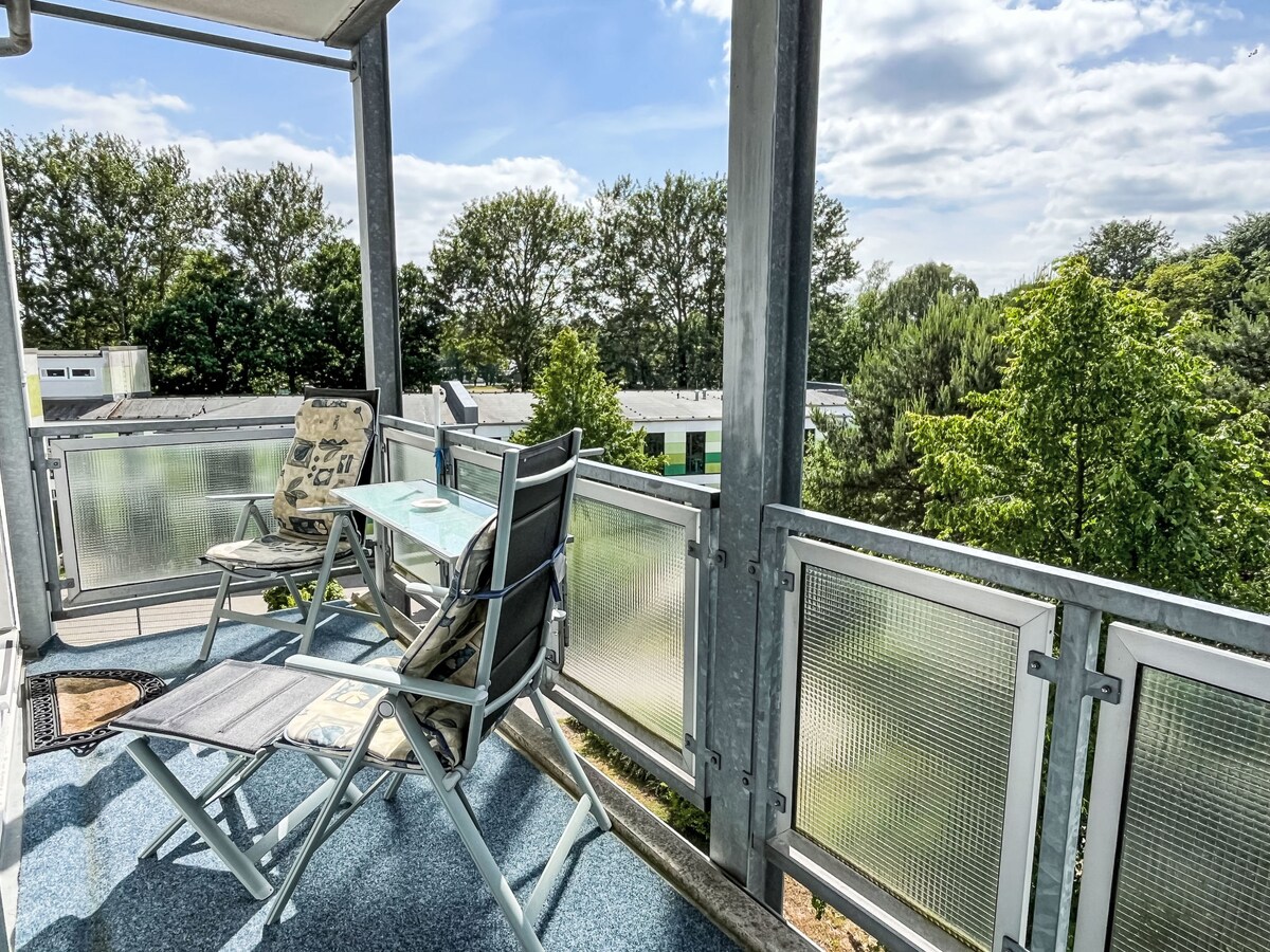 The balcony area features two compact chairs and a small table, all set against a backdrop of greenery. Sunlight filters through the trees, creating a serene outdoor space, ideal for relaxation or enjoying a meal outdoors.