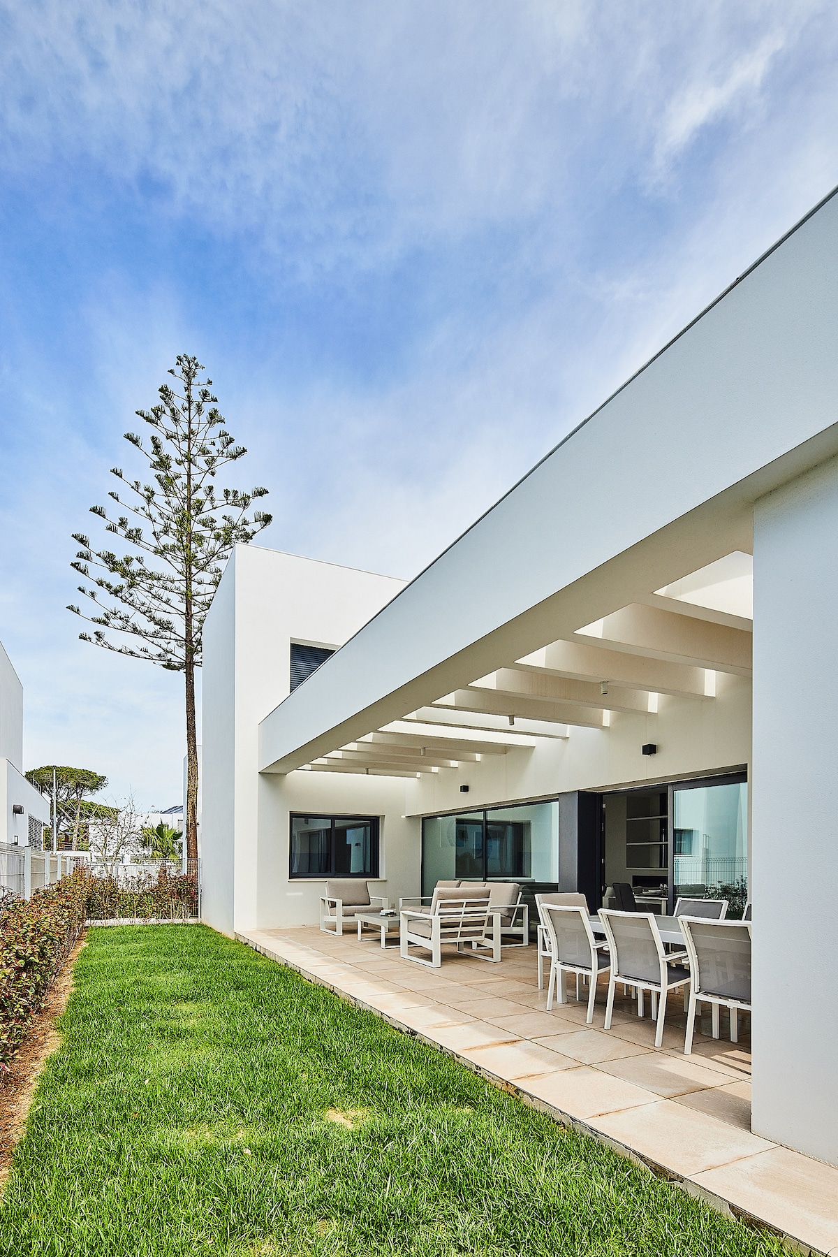 A spacious outdoor seating area is shown, featuring multiple white chairs and tables under a covered patio. Lush green grass surrounds the area, complemented by a tall tree in the background. Large windows provide a view into the airy interior of the villa.