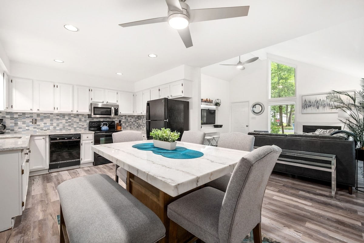 A spacious kitchen area is highlighted by a large dining table surrounded by gray chairs. Sleek countertops and modern appliances complement the bright cabinetry, while natural light pours in through large windows, creating an inviting atmosphere for meals and gatherings.