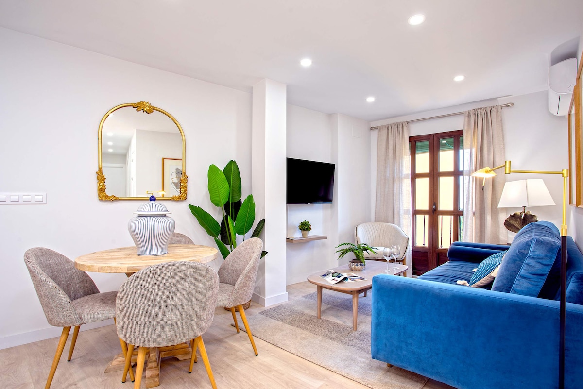 A modern open-plan living area features a round dining table with four upholstered chairs beside a cozy blue sofa. A decorative mirror hangs on the wall, and greenery adds a refreshing touch. Warm light fills the space, enhancing its inviting ambiance.