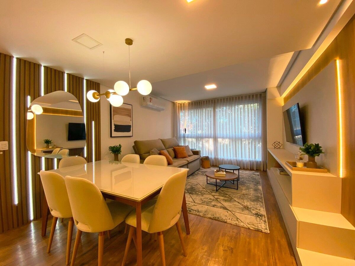 A stylish living area features a comfortable sofa, a decorative rug, and a dining table with six chairs. Natural light is allowed in through large windows, and a minimalist entertainment unit holds a flat-screen TV. Accent lighting highlights the warm tones of the space.