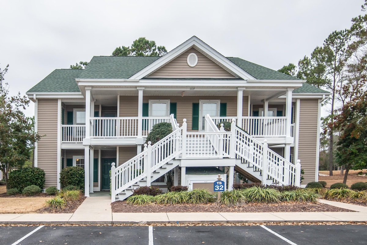 A two-story building features a light brown exterior with white trim and a green roof. Stairs lead to a second-floor entrance, flanked by vibrant greenery and neatly maintained landscaping. The building number is displayed prominently on a sign near the ground level.