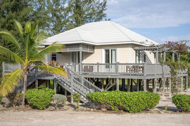 A cottage with a light-colored exterior is elevated on stilts, showcasing a spacious deck. Lush greenery surrounds the property, while palm trees and flowering plants add to the natural ambiance. Large windows are present, allowing for ample natural light.