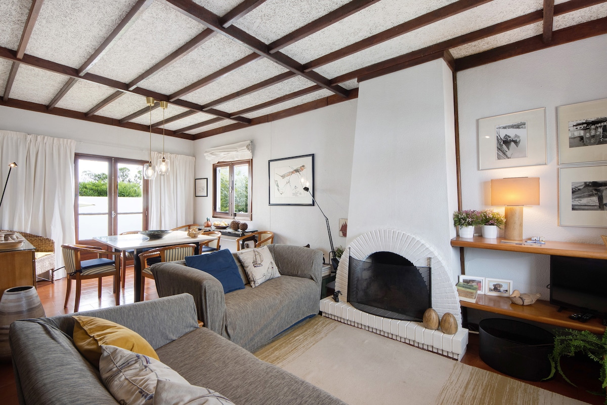 A cozy living area features a mix of comfortable seating on a light gray sofa and a warm, neutral-tone armchair. A rustic fireplace is set against a textured wall, accompanied by framed art. Natural light floods the space through large windows, adorned with soft, sheer curtains.