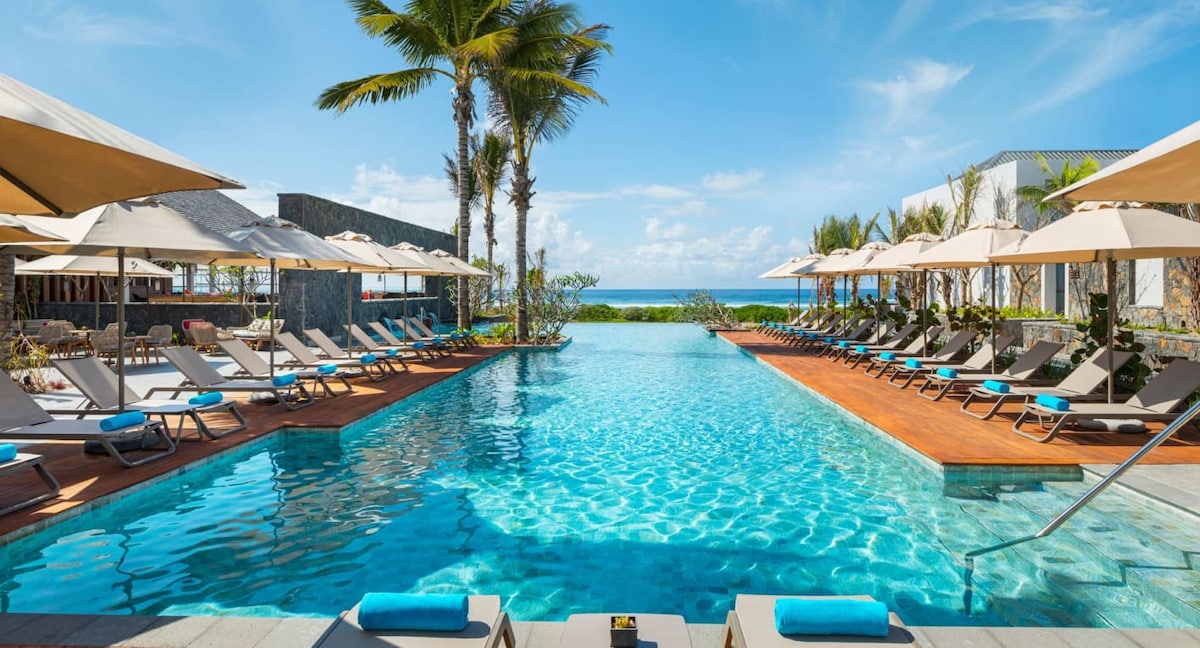 The outdoor swimming pool is surrounded by lounge chairs with umbrellas for shade. Palm trees are scattered around the area, with a clear view of the sea in the distance. The pool’s shimmering water reflects the bright sky above.