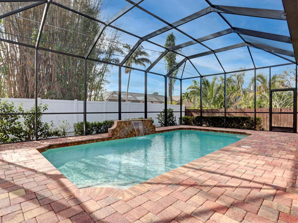 A screened-in lanai houses a heated pool, surrounded by tropical landscaping. The pool features a stone waterfall and is bordered by a brick patio. The area is enclosed, providing a sense of privacy while allowing for views of greenery beyond.