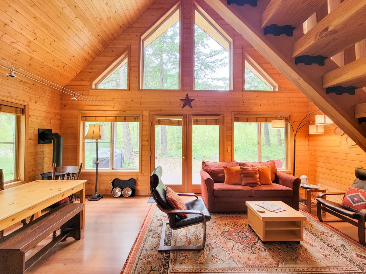 The interior of the cabin showcases a warm wood-paneled living area with tall windows allowing natural light to fill the space. A cozy couch with accent pillows sits next to a coffee table, while a dining table is positioned nearby. A cast iron stove is also visible.