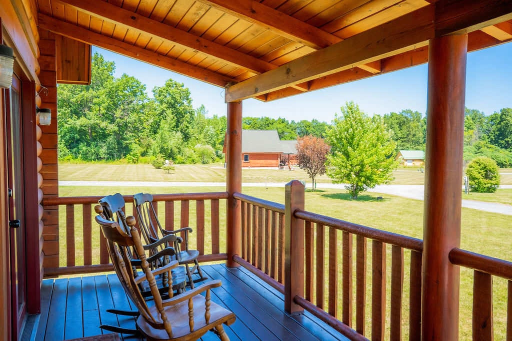 A spacious front porch features rocking chairs positioned along the railing, inviting relaxation with scenic views of the surrounding greenery and open space. Sunlight streams down, enhancing the charming wooden structure and its inviting atmosphere.