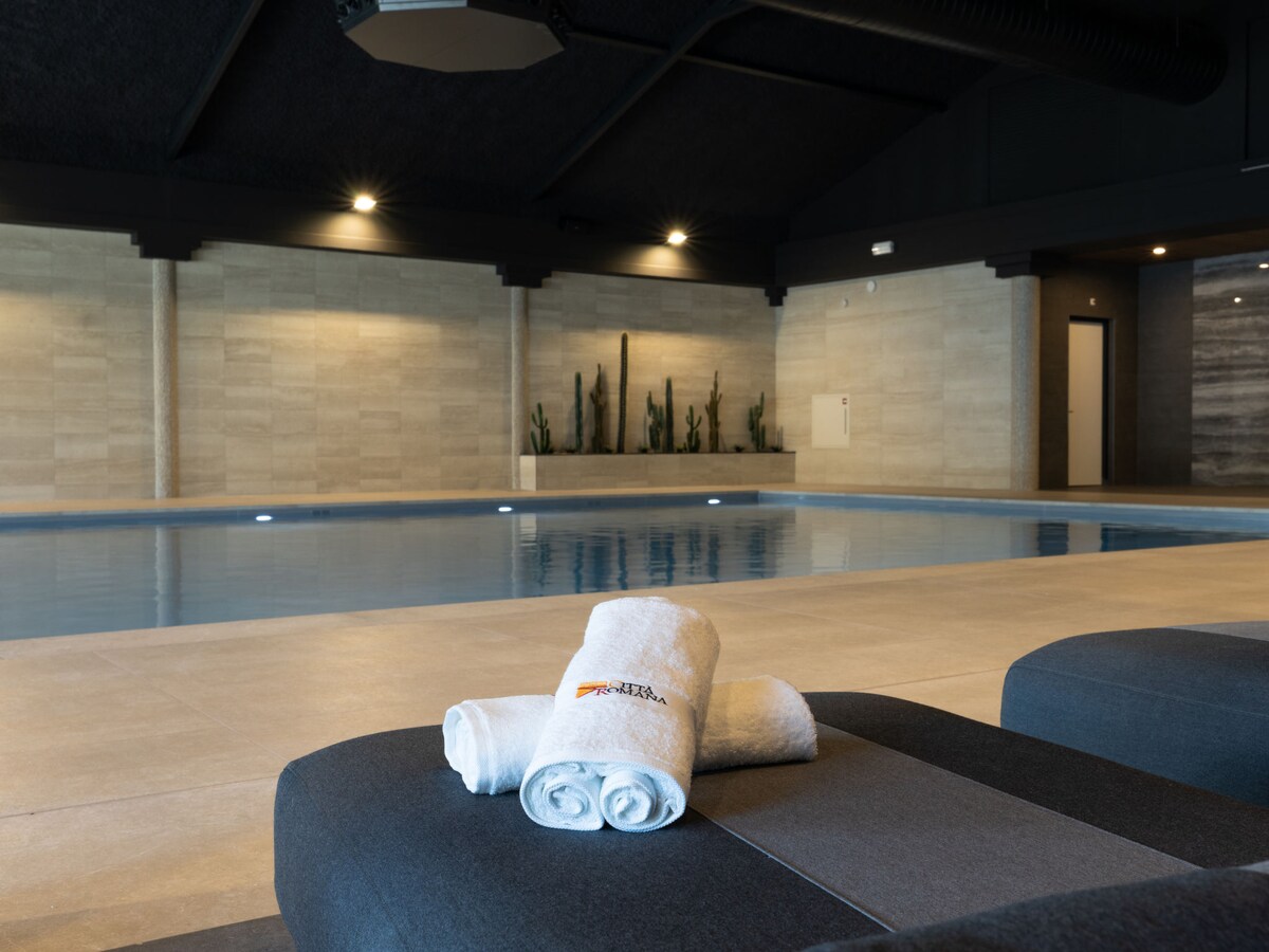 An indoor pool area is featured, with a serene water surface reflecting the soft lighting. Grey-toned tiles and minimalist decor enhance the modern aesthetic. Plush towels are neatly arranged on a seating area, with decorative cacti subtly positioned in the background for a touch of nature.