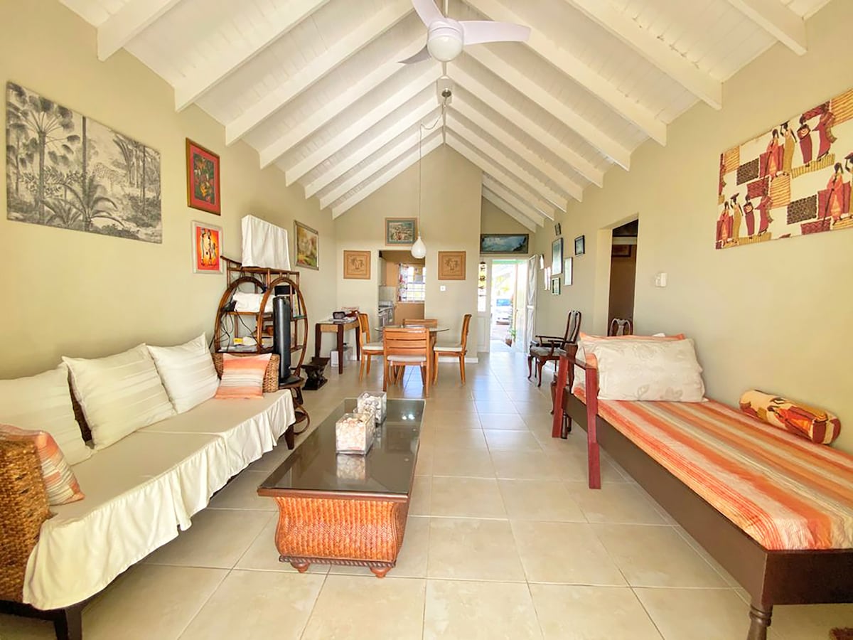 An open-plan living and dining area features a high ceiling with wooden beams. A comfortable seating arrangement includes a sofa and armchairs. Decorated walls display various art pieces. A wooden dining table is visible, with natural light entering through the patio doors.