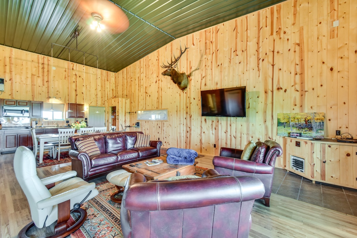 A spacious living area features wood-paneled walls and high ceilings, offering a cozy yet open feel. A mounted deer head adds a rustic touch. Leather sofas and armchairs provide comfortable seating, complemented by a wooden coffee table and television mounted on the wall.