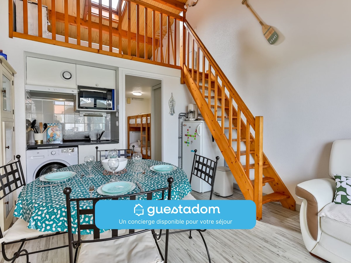 The image captures a bright living area with a round dining table set for four. The kitchen is visible, equipped with modern appliances. A wooden staircase leads to the mezzanine sleeping area. Light streams in through a window, enhancing the open feel of the space.