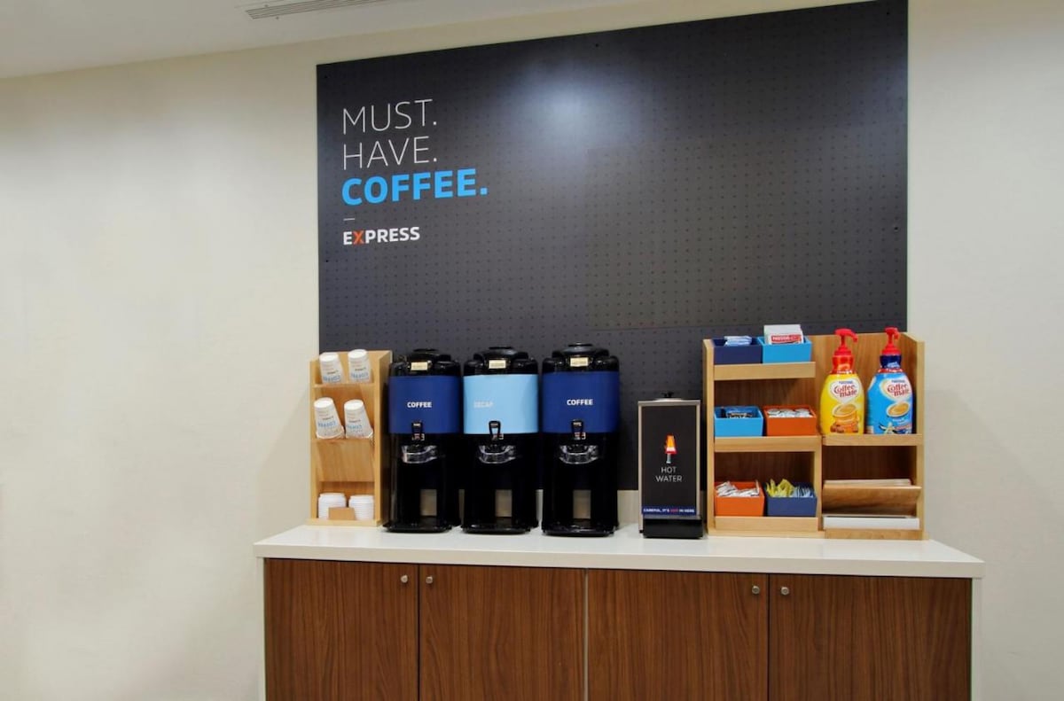 A coffee station is presented with two coffee dispensers and stacks of disposable cups. Shelf displays include coffee creamer options and sweeteners, all arranged on a countertop beneath a wall sign that reads, 'MUST. HAVE. COFFEE.'
