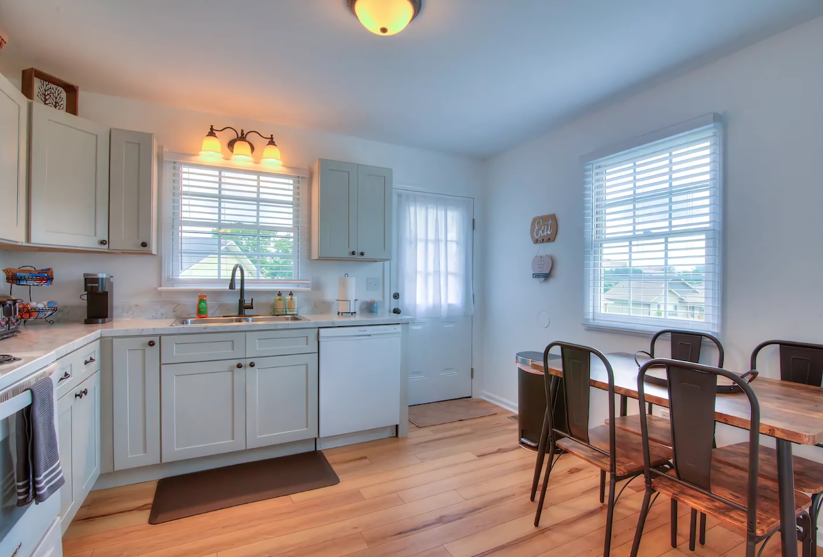 The kitchen features modern cabinetry and ample countertop space, illuminated by natural light from the windows. A dining table with seating for four is positioned nearby, creating a functional space for meals. The appliances are designed for ease of use, enhancing the cooking experience.