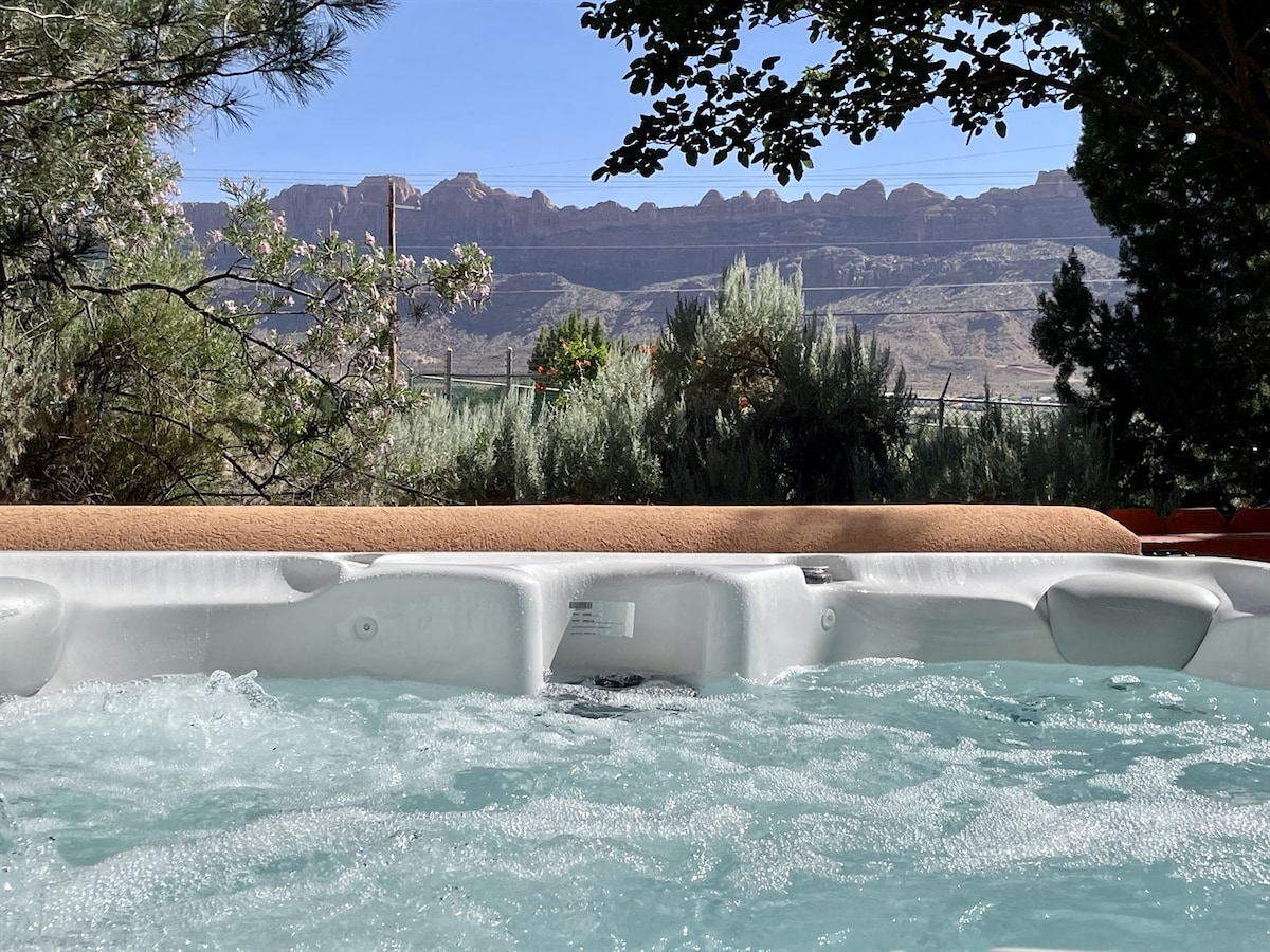 A private hot tub is seen bubbling with water amidst a serene outdoor setting. In the background, the stunning red rock formations of Moab create a dramatic landscape under a clear blue sky, enhancing the feeling of relaxation and seclusion.