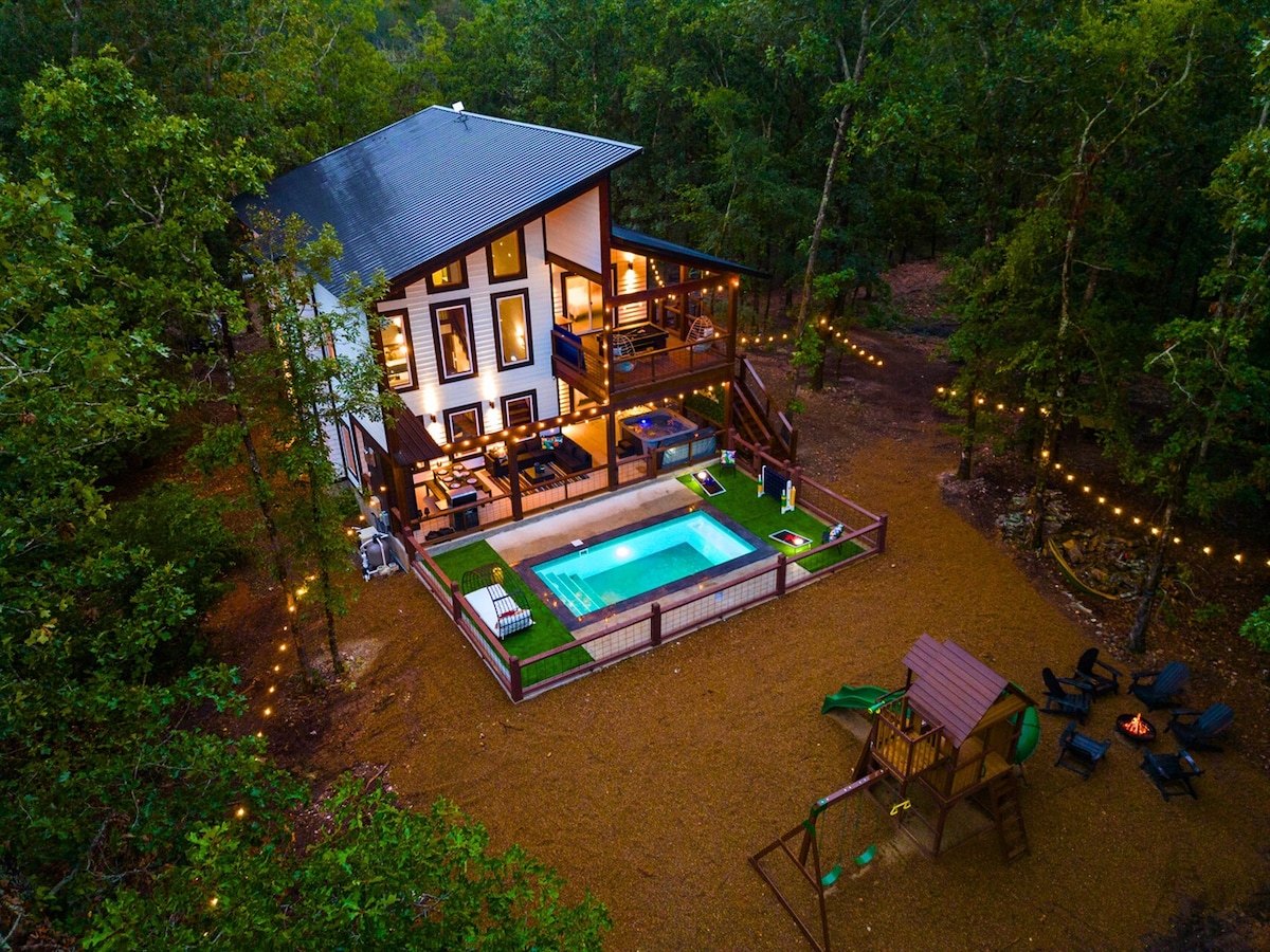 An aerial view showcases a modern three-story cabin surrounded by lush trees, featuring a private heated pool enclosed by a wooden fence. A playset with swings stands in the yard, and warm lights create an inviting atmosphere around the patio and fire pit area.
