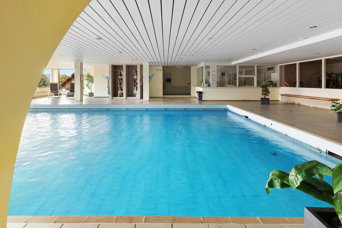 An indoor pool is featured, with clear blue water reflecting light from the ceiling. Surrounding areas include tiled flooring and windows that provide natural light. Lounge chairs are placed nearby, and potted plants add a touch of greenery to the space.