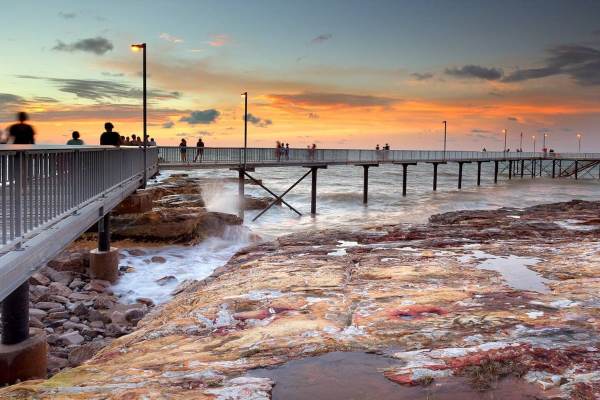 A wooden pier extends into the water, with people walking along its path. The scene is illuminated by soft lighting and features a vibrant sunset with hues of orange and purple reflected on the water. Rocky shoreline frames the left side of the image.