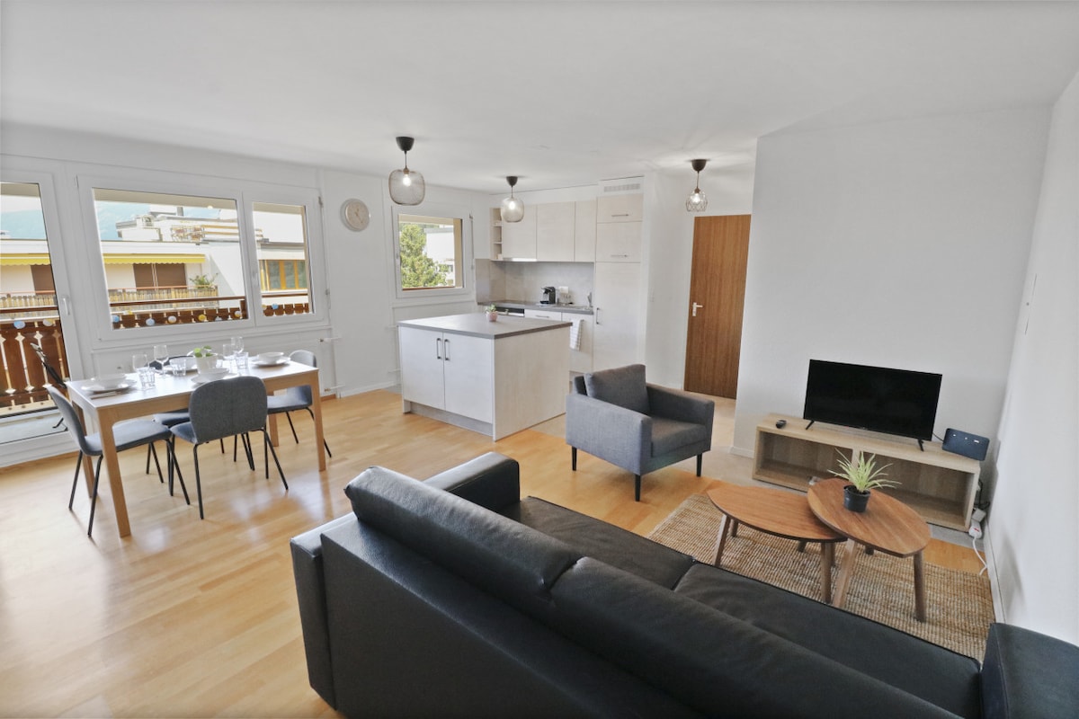 A spacious living area features a modern open kitchen with light cabinetry and a dining table set for four. Comfortable seating is arranged around a wooden coffee table, while large windows allow for natural light to fill the space.