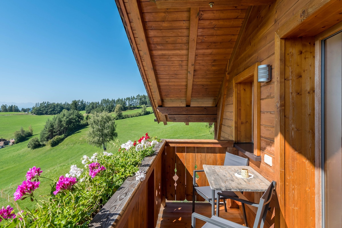 A wooden balcony features a small table with two chairs, surrounded by vibrant pink and white flowers. Lush greenery and distant hills can be seen beyond, with clear blue skies overhead, creating an open and inviting outdoor space.