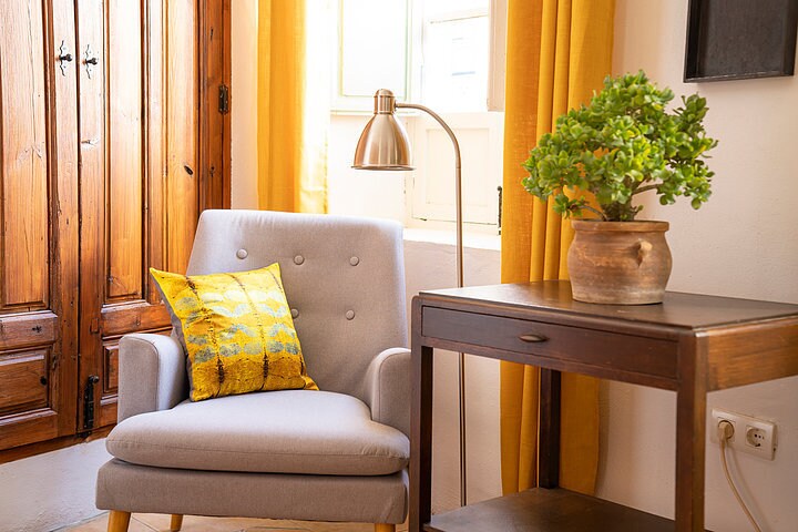 A cozy corner is highlighted by a light gray armchair with a yellow decorative pillow. A small wooden side table stands beside the chair, supporting a potted plant. Warm yellow curtains frame a window, allowing natural light to brighten the space.