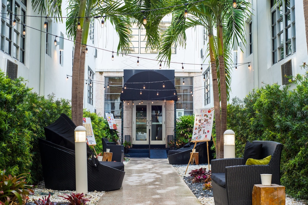 A welcoming courtyard is flanked by lush greenery and swaying palm trees. Cozy seating areas are provided with dark wicker chairs and side tables, while string lights create a charming ambiance. The main entrance to the building features large windows and an inviting black awning.