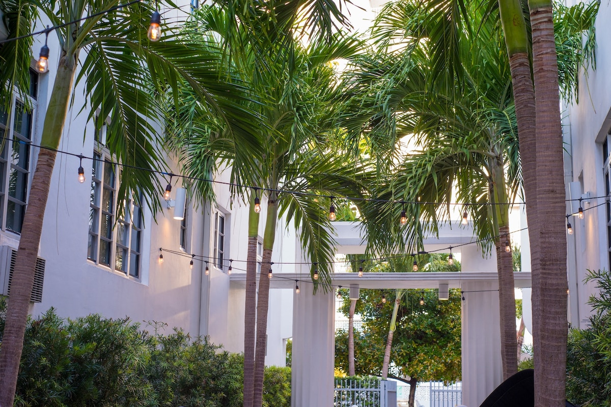 A serene outdoor area is lined with tall palm trees and lush greenery. Soft string lights are draped overhead, creating a warm ambiance. The backdrop features a sleek white structure that frames the space, inviting guests to enjoy the tropical surroundings.