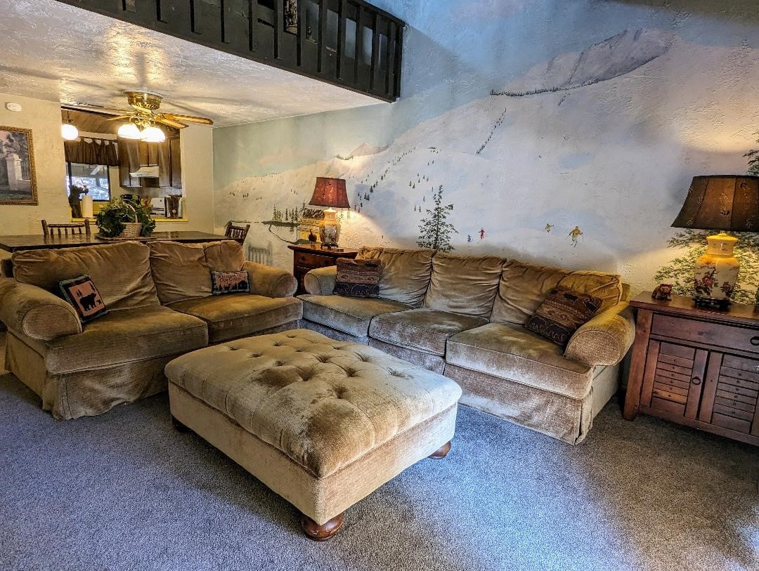 A spacious living room is furnished with a large sectional sofa and a tufted ottoman. Soft lighting is provided by two lamps, creating a cozy ambiance. A mural of a snowy mountain scene with trees adds a serene backdrop to the space.