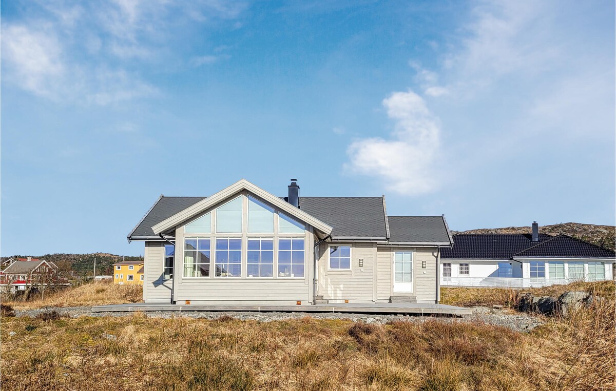 A spacious cottage exterior is presented against a clear blue sky. Large panoramic windows reflect the natural landscape, while a wooden deck extends towards the landscaped grounds. The light-colored building features a gabled roof, harmonizing with the serene surroundings of the area.