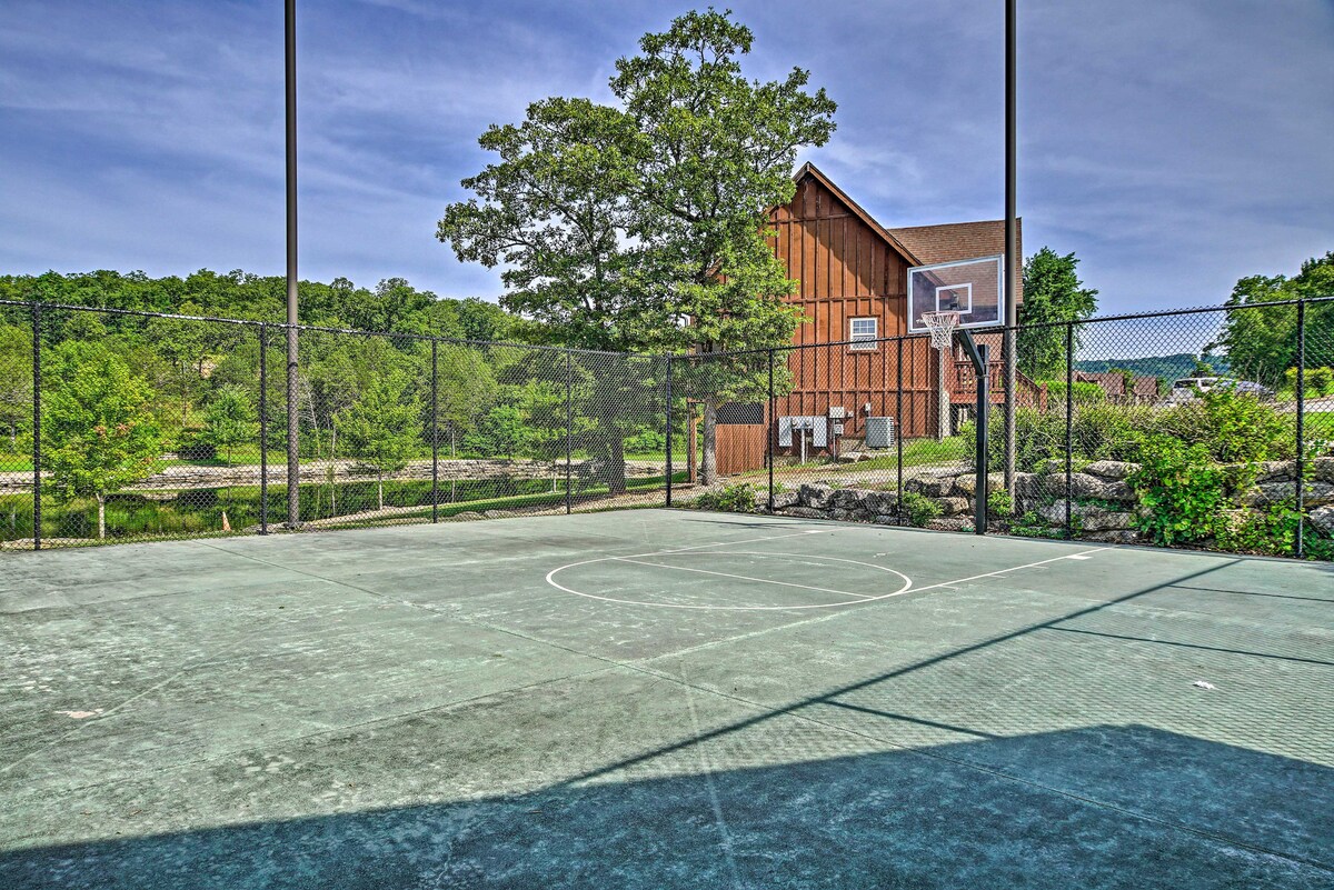 A basketball court is enclosed by a chain-link fence, featuring a smooth surface marked with a half-court line. In the background, a wooden building is visible amidst green trees, while a fishing pond is located just beyond the court.