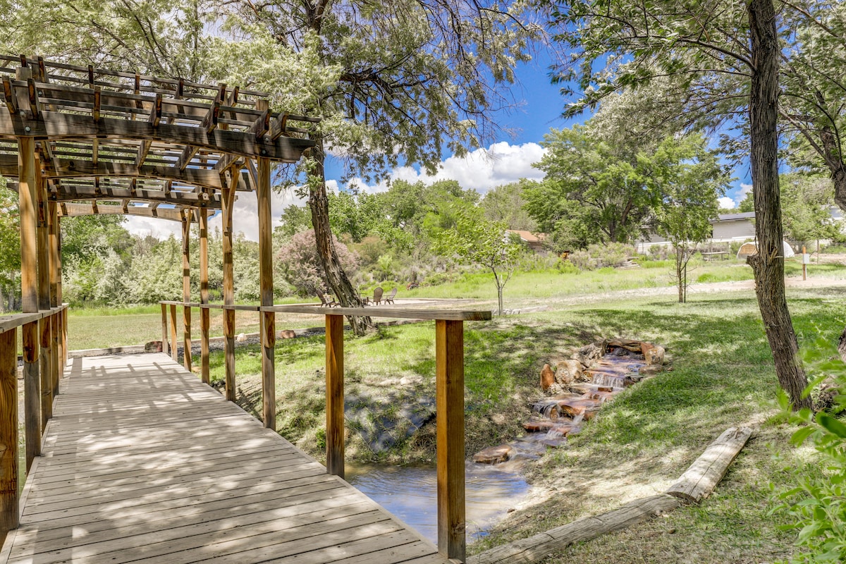 A wooden pedestrian bridge is seen leading over a gentle creek, surrounded by lush greenery and trees. The clear blue sky provides a bright backdrop, enhancing the natural beauty of the landscape, which features a grassy expanse and distant hills.