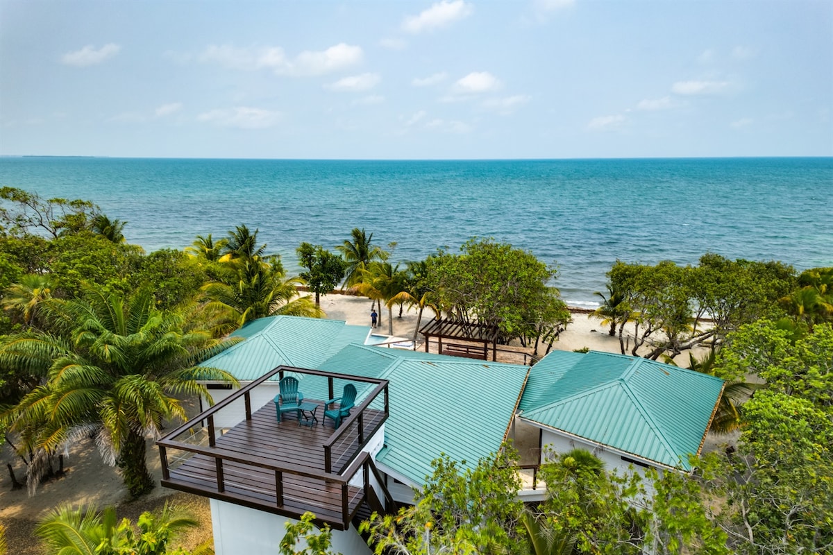 An aerial view captures the beachfront property, featuring a rooftop deck with seating overlooking the turquoise ocean. Lush greenery surrounds the structures, and the sandy beach is visible in the distance. The building features multiple roofs with a distinct green color blending into the landscape.