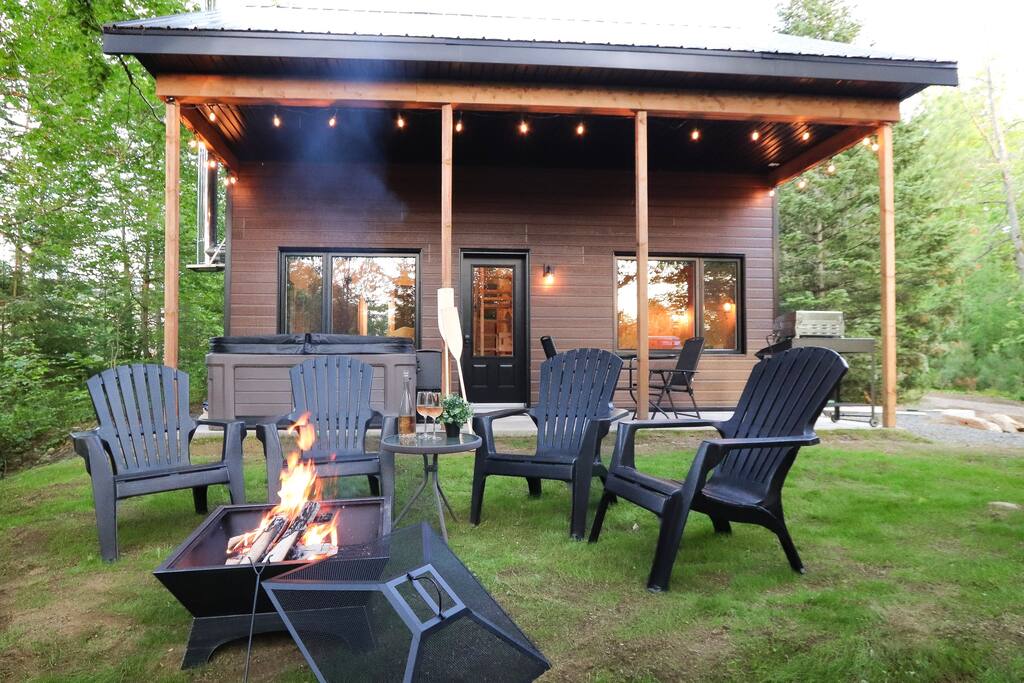 An outdoor seating area is presented with five black Adirondack chairs arranged around a fire pit. A covered porch extends from the cabin, featuring warm lighting and large windows that invite natural light. A hot tub is located to the side, surrounded by greenery.