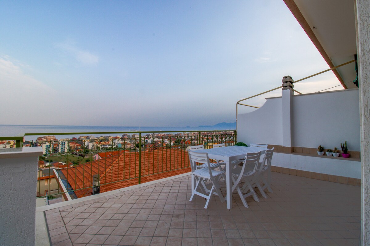 A spacious terrace features a white outdoor dining set. A scenic view captures the coastline and rooftops of the neighborhood. The clear sky transitions to soft hues, enhancing the serene atmosphere. Potted plants are placed along the terrace wall, contributing to the inviting outdoor space.