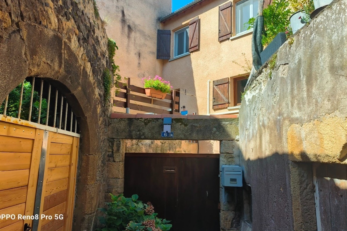 The exterior of the house is depicted, showcasing a charming balcony adorned with potted flowers. A stone wall and wooden gates frame the entrance, while the structure features beige stucco and shuttered windows. A mailbox is visible beside the garage door.