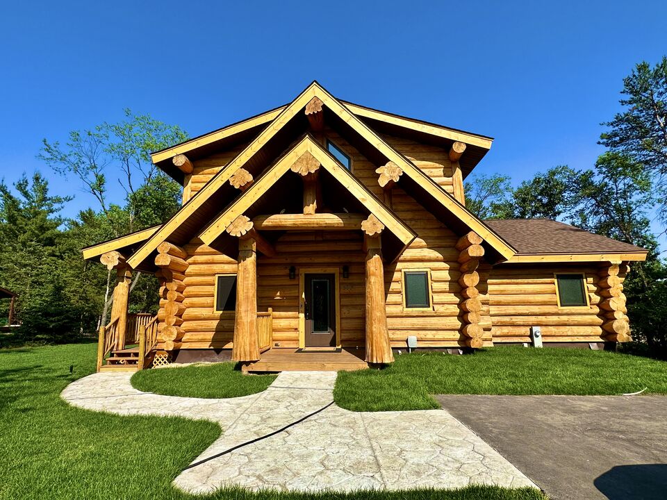 The log home features a rustic exterior with prominent wooden beams and decorative details. A spacious front porch is accessible by a winding concrete pathway, framed by lush green grass and trees in the background offering a natural setting.